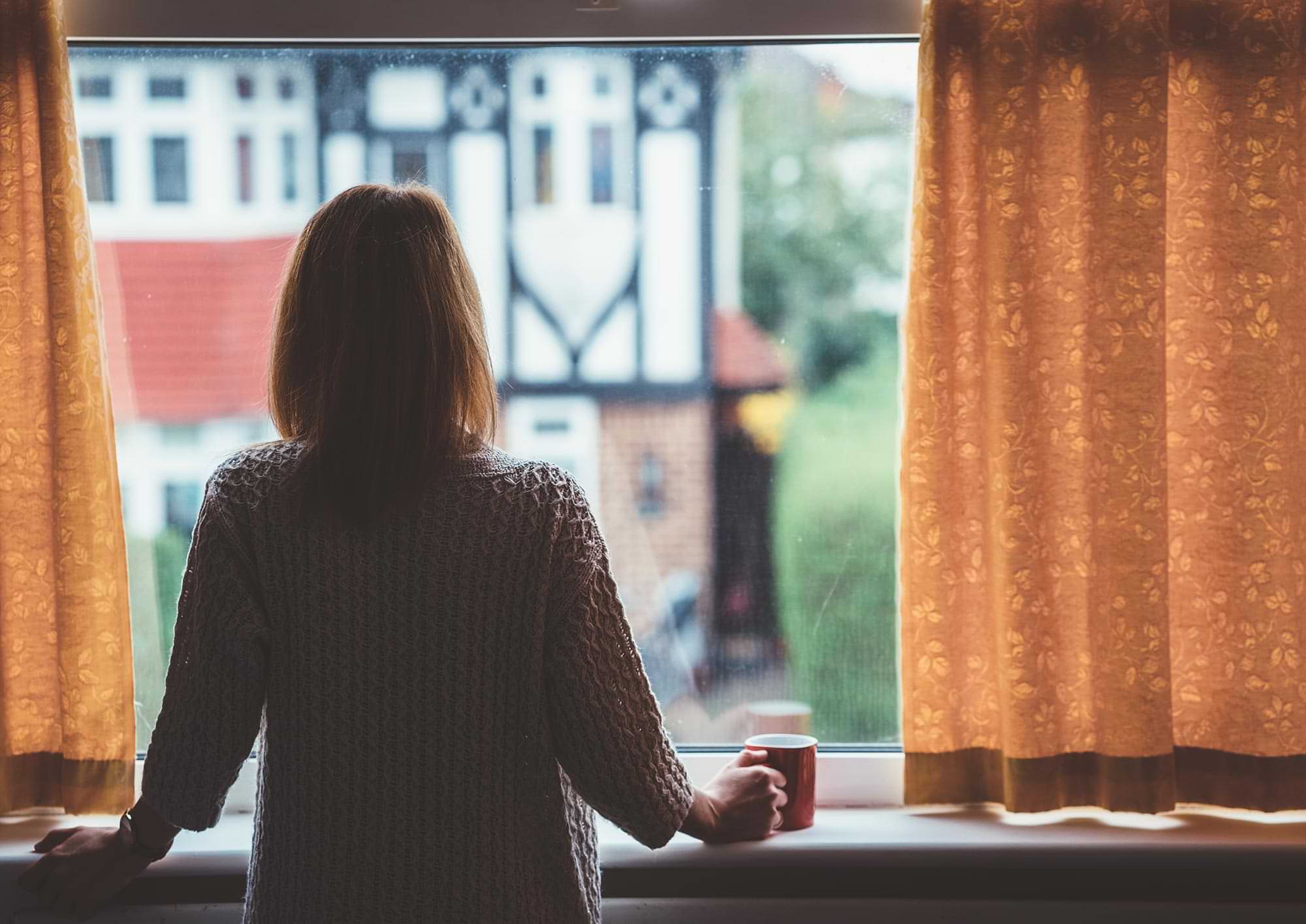 back view of a woman looking out a window, grasping a mug in one hand