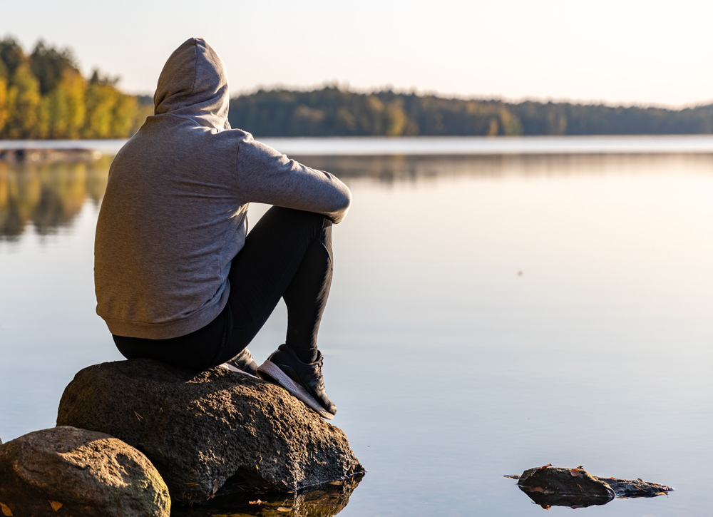 man with hood on sitting on rock in lake