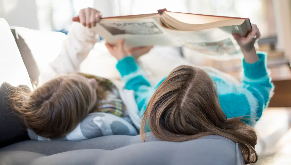 two girls reading a book together while lying down on the couch