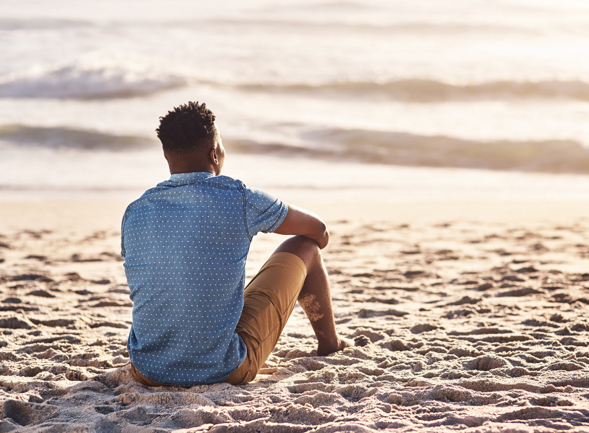 view of man's back while he's sitting in the sand at a beach