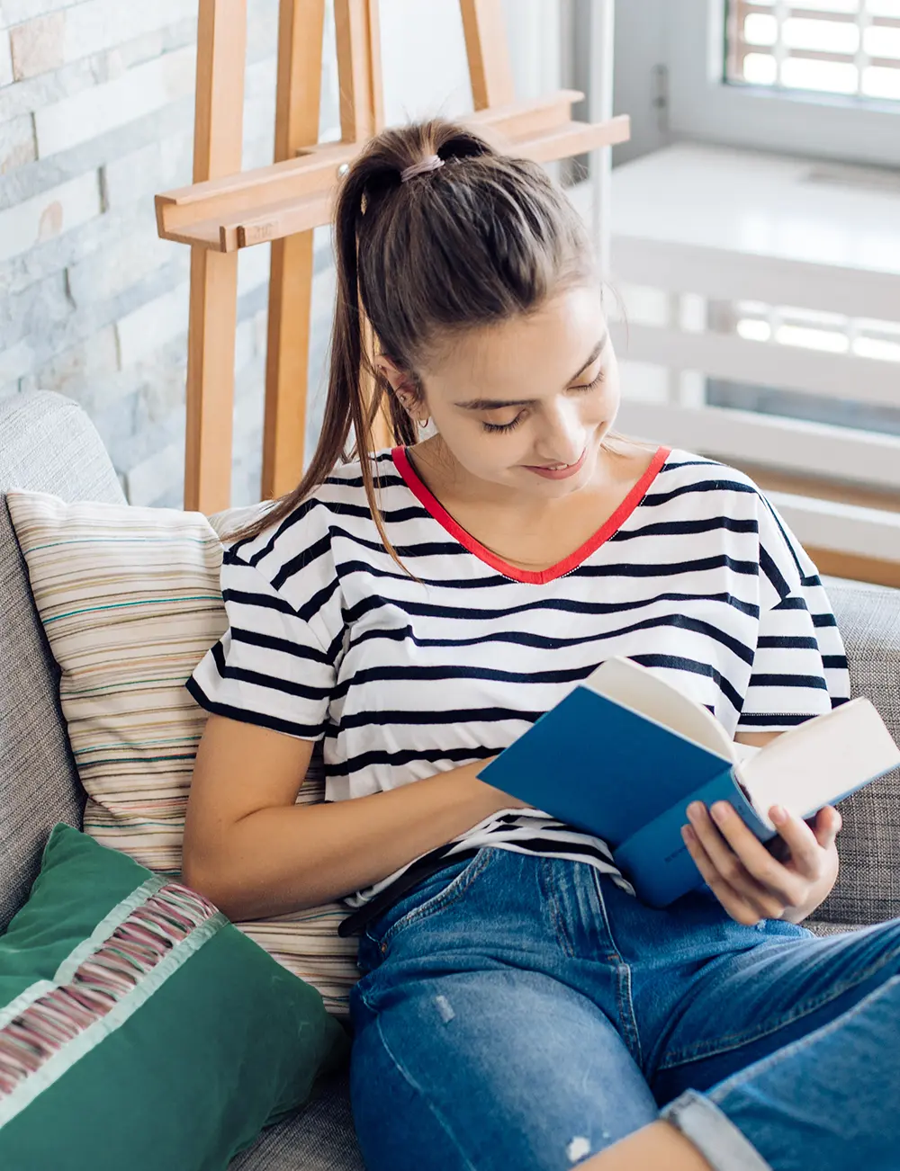 Digital stock image photograph display cropped portrait orientation view of a young woman with a dark brown high ponytail seated on a couch and reading a book with a blue cover; She is wearing a black and white striped short sleeved t-shirt that contains a red colored v-neck line and blue denim jeans as she is smiling while looking down at the book pages