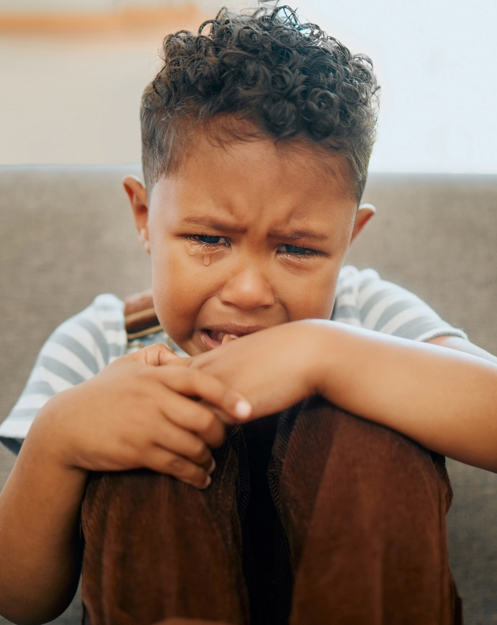 A young child with curly hair crying, wearing a striped shirt and brown overalls, sitting with their hands over their knees indoors.