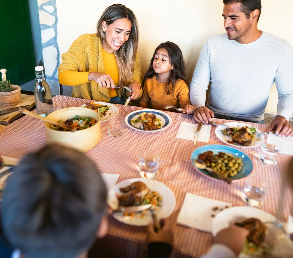 Family of four seated around a dining table covered with a red and white checkered cloth, enjoying a home-cooked meal together.