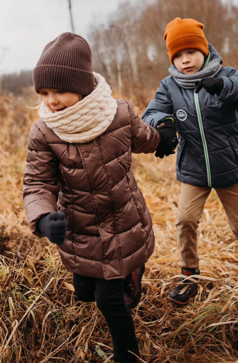 Two young children, bundled in winter coats, scarves, and hats, running through a dry, grassy field while holding hands.