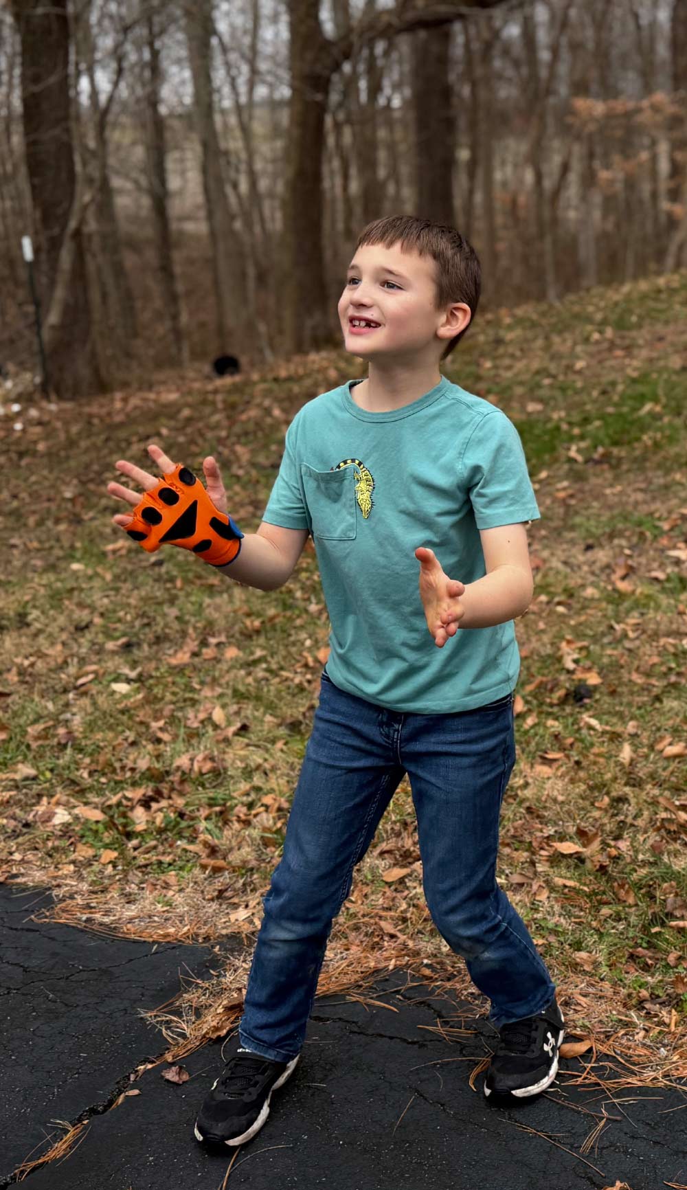 A young boy in a teal shirt and jeans stands outdoors on a paved surface, wearing an orange and black athletic glove and looking upward.