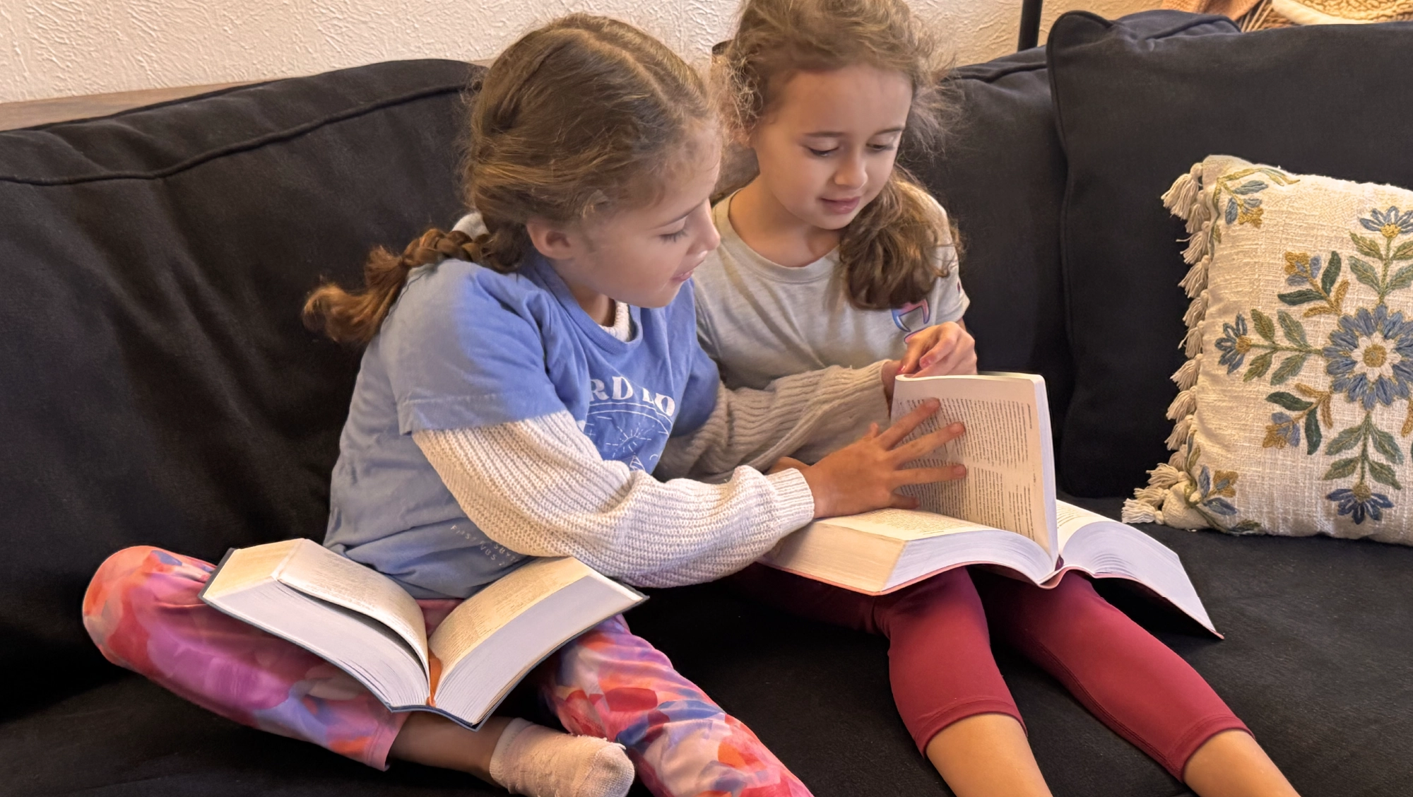 Two young girls sit on a dark couch together, each holding an open book and looking at the pages