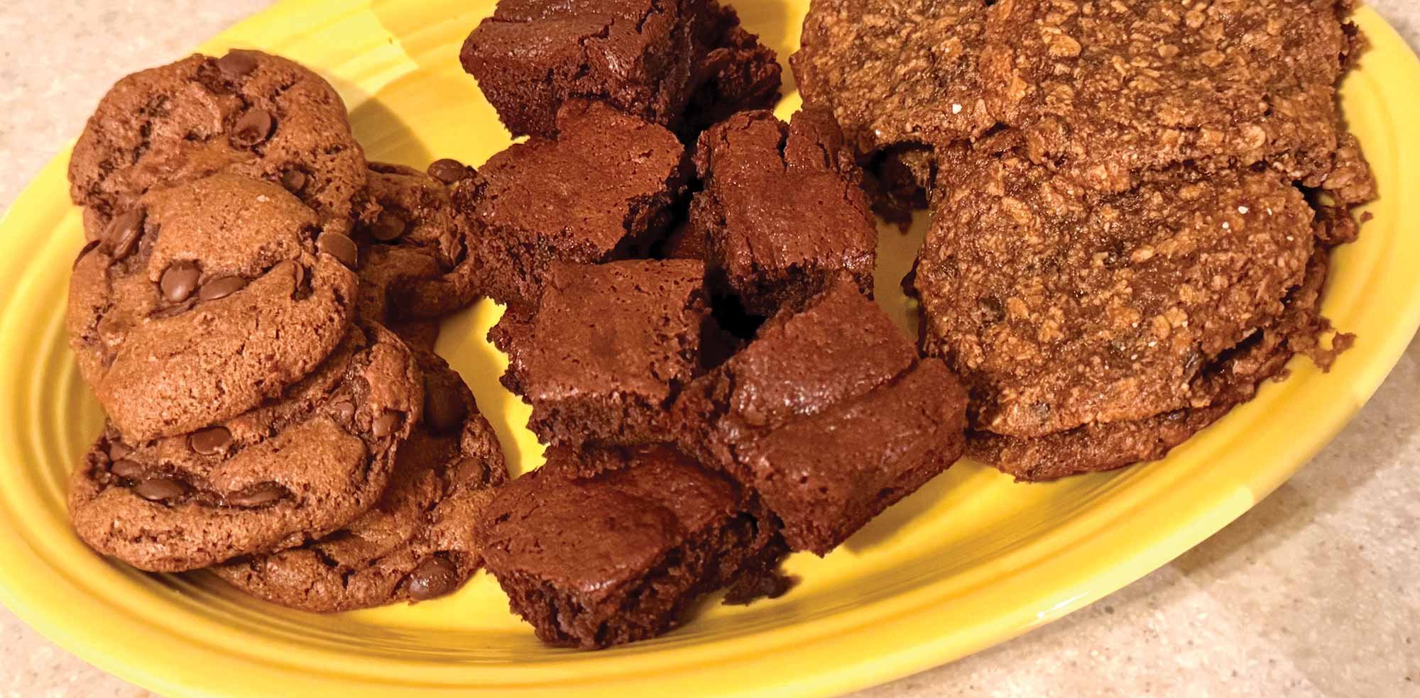 An assortment of freshly baked chocolate chip cookies, square brownie bites, and oatmeal cookies served on a yellow platter.