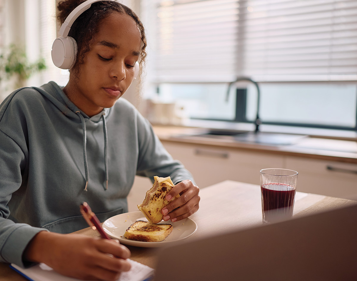 A teenage girl wearing headphones writes in a notebook while eating breakfast at a table.