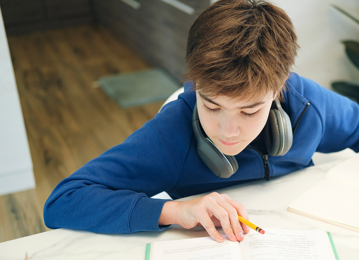 A young boy with headphones around his neck leans forward over an open textbook.