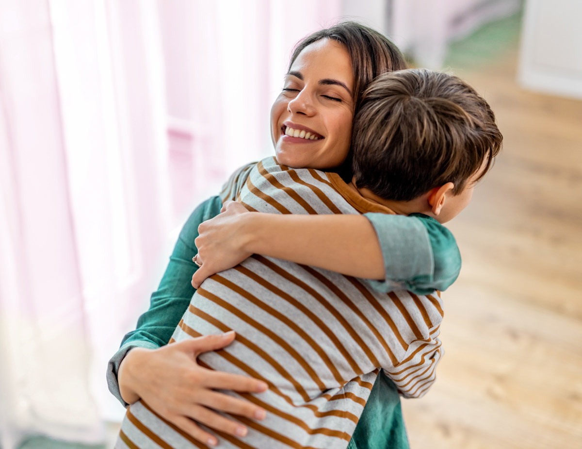 A smiling mother closes her eyes and happily embraces her young son in a warm hug inside their home.