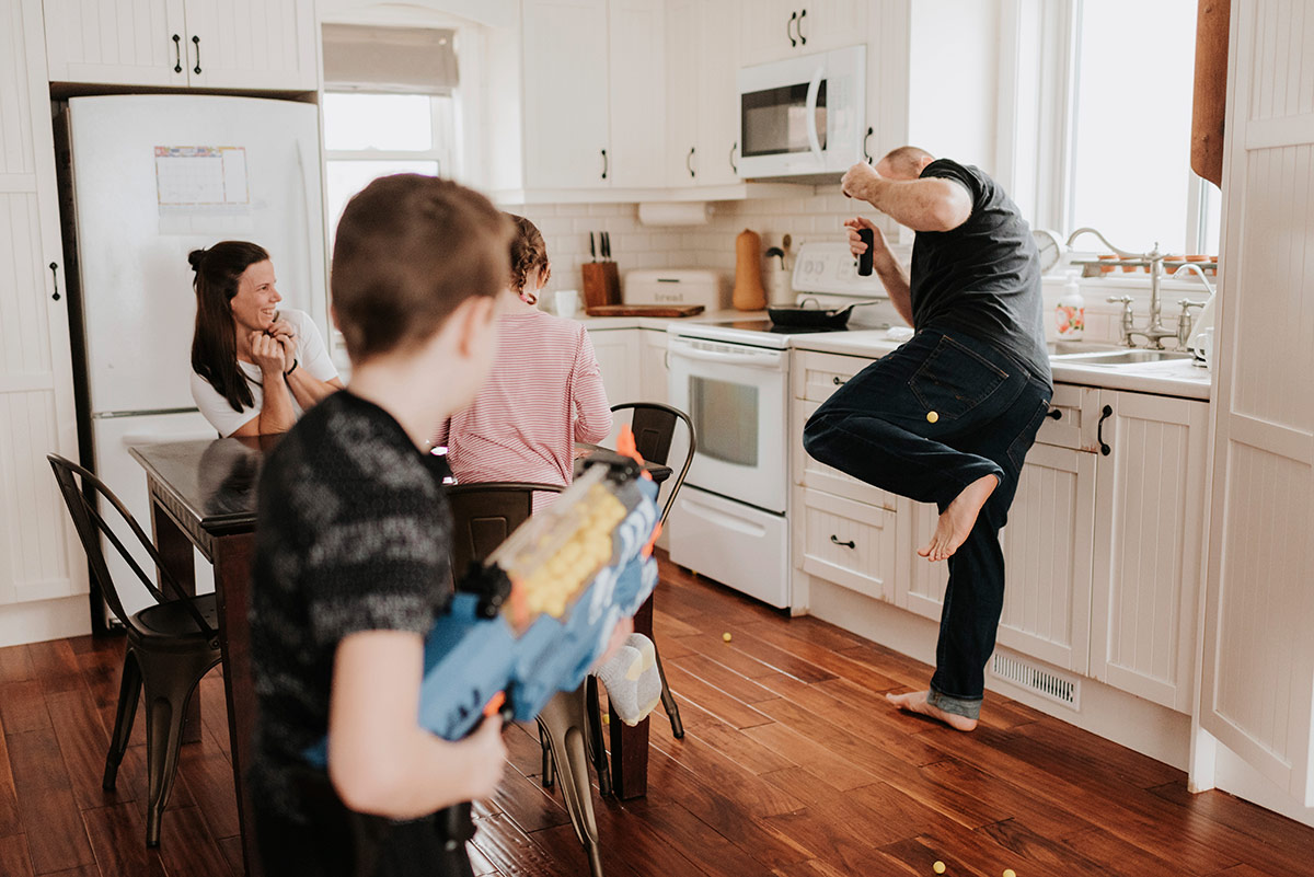 A family plays a lively toy blaster game in a bright kitchen, with a dad dodging foam darts while the mom laughs from the table.