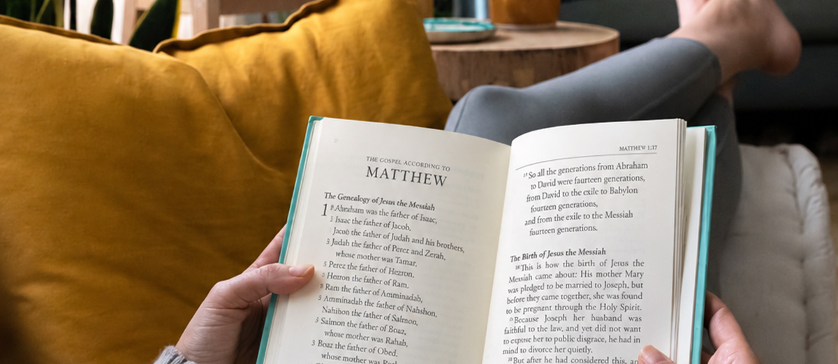 A person holds open a book to a page titled "THE GOSPEL ACCORDING TO MATTHEW." Yellow cushions are visible on the left, and the person's feet rest on a cushion in the blurred background.
