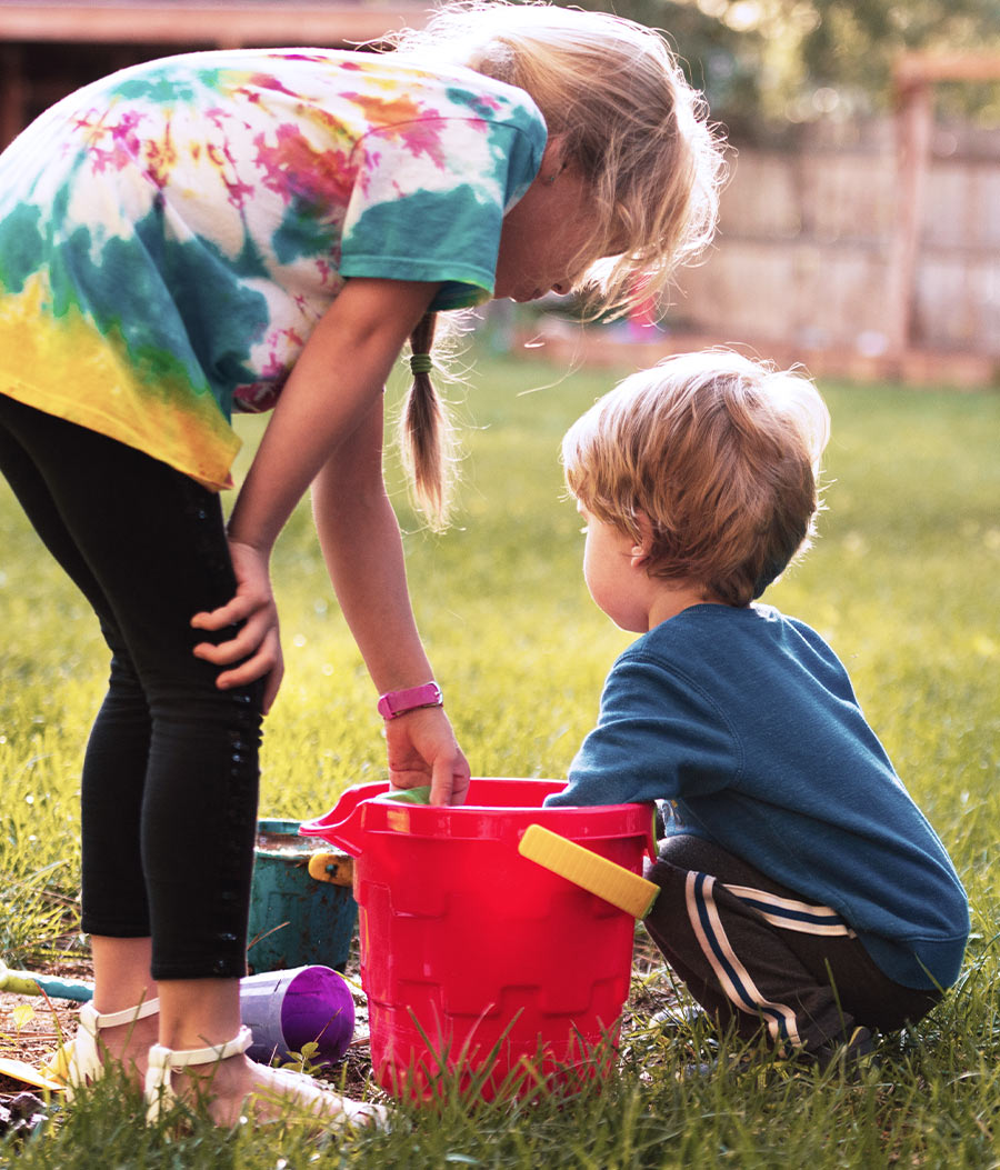 A young girl in a tie-dye shirt stands on a grassy lawn, leaning down toward a young boy who is crouching next to a large red plastic bucket.