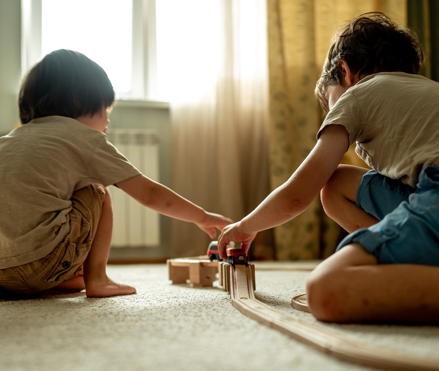 Two young boys crouch on a carpeted floor, playing together with a wooden toy train track assembly in front of a sunlit window.
