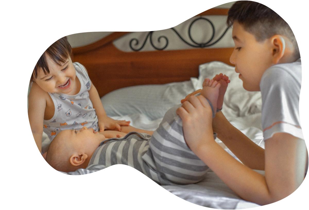 An older boy holds a baby's feet while lying on a bed next to a smiling toddler who looks down at the baby.