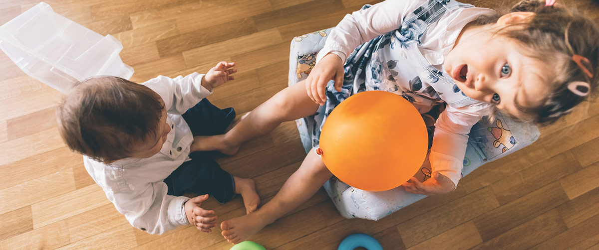 An overhead view of two young children playing on a wooden floor, with one holding a large orange balloon.
