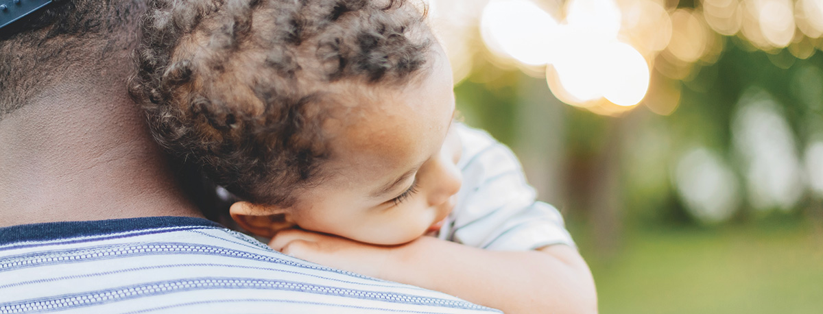 A young child rests their head gently on a person's shoulder outdoors.