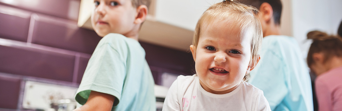A toddler smiles at the camera with other young children playing nearby.