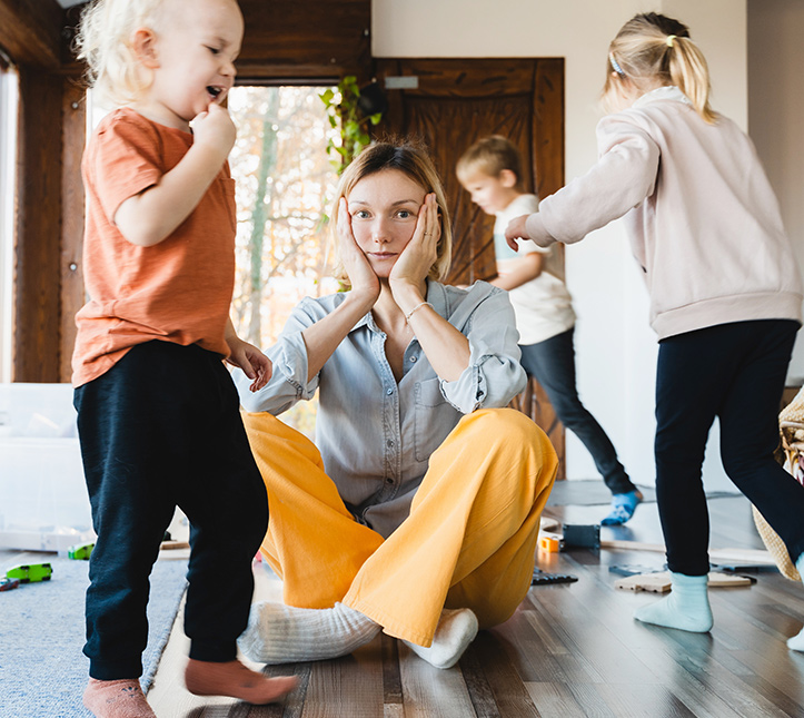 A stressed mother sits on a living room floor surrounded by active, playing children.