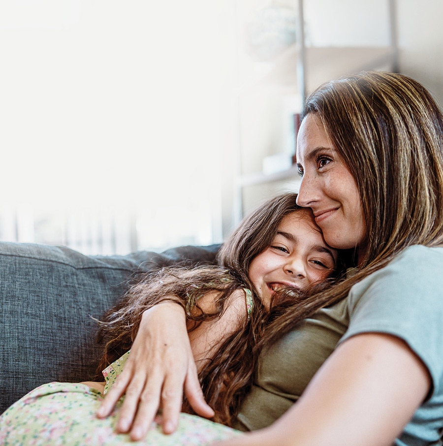 A smiling mother cuddles with her young daughter on a couch at home.