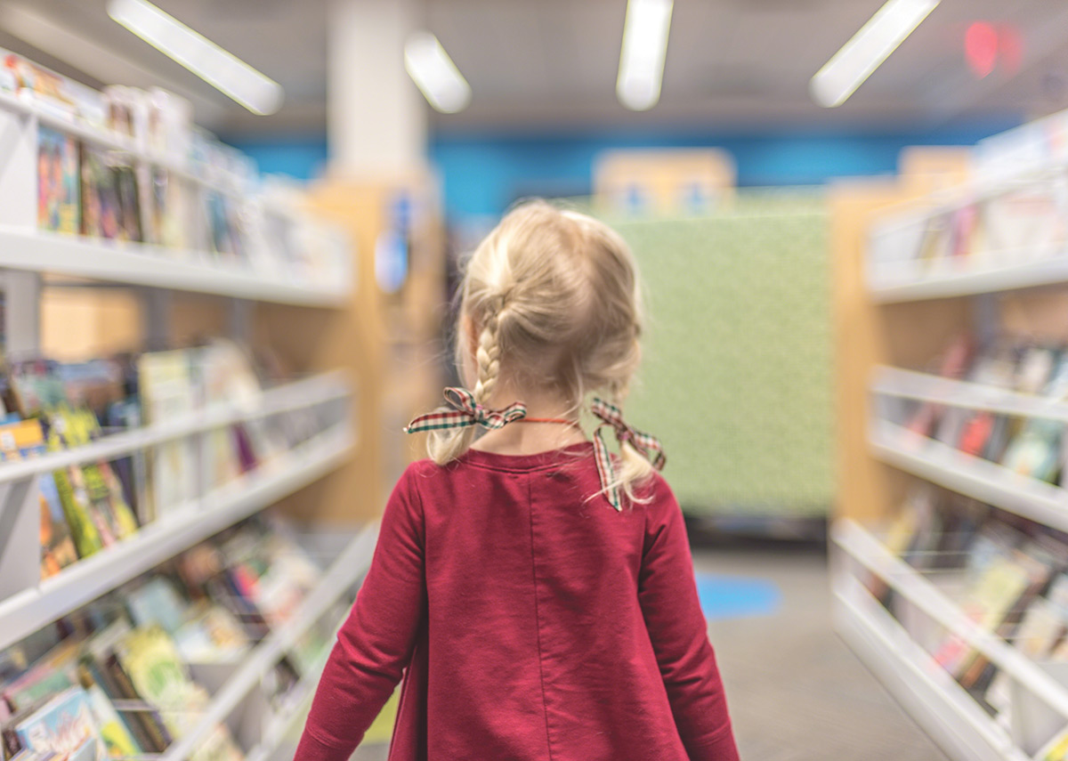 Back view of a young girl with braided hair walking down a library aisle.