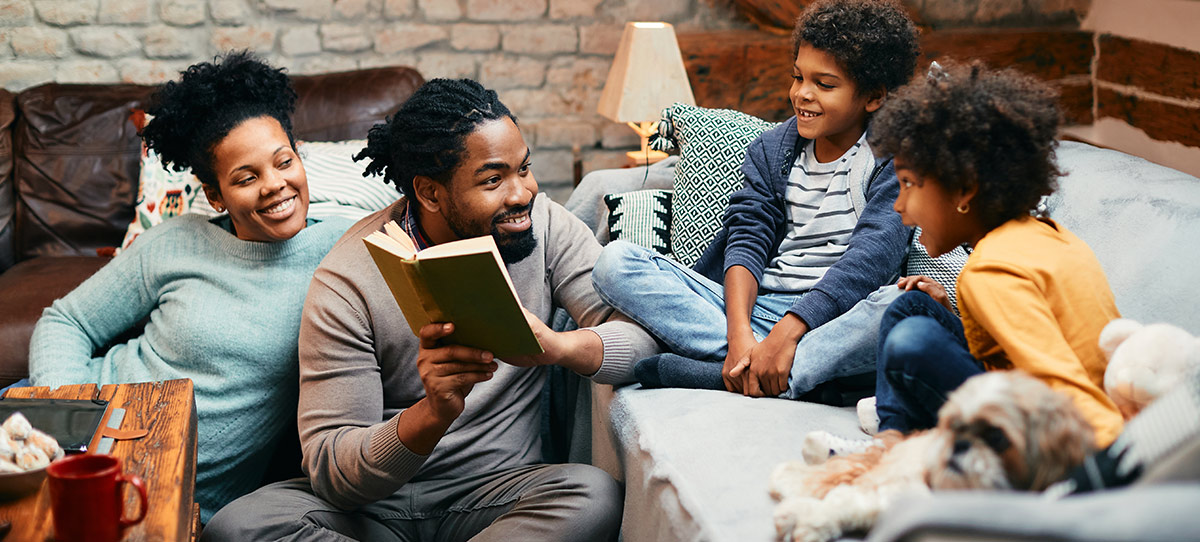 A father sitting on the floor reading a book aloud to his smiling wife and two children gathered on the sofa in a cozy living room