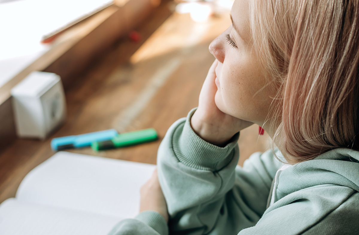 Profile view of a young blonde girl in a mint sweatshirt resting her chin in her hand while seated at a desk.